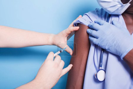 Photo for African american nurse wearing face mask, getting vaccinated with syringe and needle. Close-up of medical assistant receiving vaccine shot from hands of doctor for virus protection. - Royalty Free Image