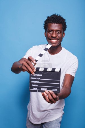 Téléchargez les photos : Le cinéaste afro-américain tenant un battant ouvert dans les mains, l'air confiant avec un sourire au visage. Producteur de film noir avec clapperboard debout fier devant fond bleu isolé. - en image libre de droit