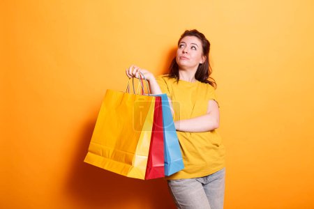 Photo for Happy, stylish brunette shopper carries shopping bags and smiles on isolated orange background. Caucasian woman looking attractive and cheerful while grasping colorful parcels. - Royalty Free Image