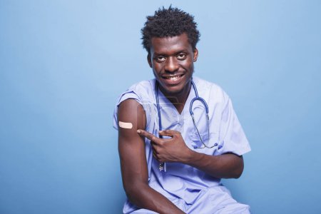 Photo for African american medical nurse having adhesive bandage after vaccination against coronavirus. Vaccinated male nurse with scrubs pointing at vaccine shot patch for immunity. - Royalty Free Image