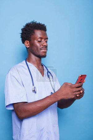 Photo for African American doctor in scrubs wears stethoscope and holds a smartphone, smiling confidently in a studio. Black modern healthcare professional utilizing mobile device during covid 19 pandemic. - Royalty Free Image