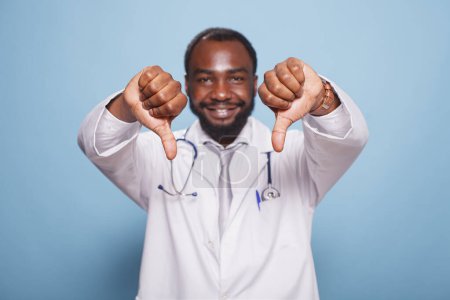 Photo for Black man wearing a hospital uniform and stethoscope standing and doing thumbs down to the camera. Portrait of african american medical specialist making disapproval hand gestures. - Royalty Free Image