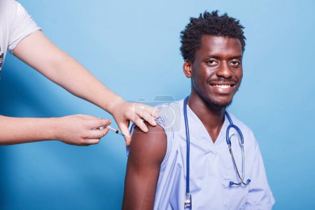 Photo for Male medical nurse wearing blue scrubs getting vaccine injection with syringe in studio. African American physician with stethoscope, getting vaccinated for coronavirus immunity. - Royalty Free Image