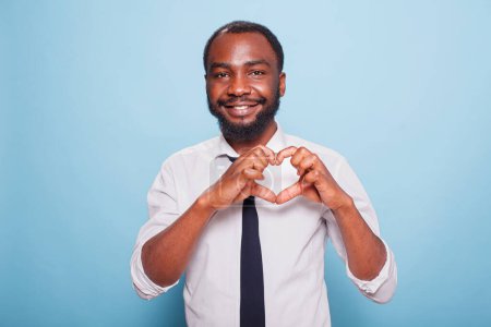 Téléchargez les photos : Sourire influenceur afro-américain faire des doigts en forme de coeur pour montrer le soin de supporter les fans. Joyeux blogueur masculin en chemise blanche et cravate noire faisant le geste de la main pour l'amour sur sa poitrine. - en image libre de droit