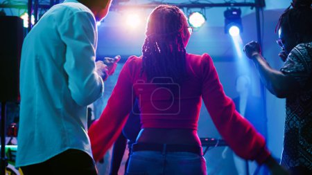 Photo for Men and women dancing at nightclub and listening to music, having fun at club with stage lights. Group of persons enjoying social gathering with friends on dance floor. Handheld shot. - Royalty Free Image