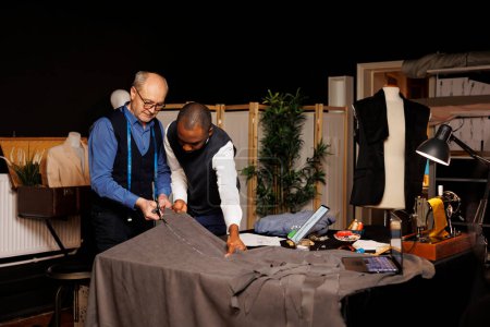 Skilled craftmanship by professional artisan menswear specialist and african american apprentice in tailoring workspace. Masterful old fashioned tailors creating elegant attire from scratch