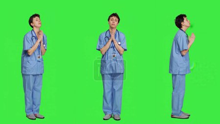 Photo for Medical assistant being hopeful praying for good luck against greenscreen, holds hands in a prayer sign and having belief in spirituality. Young christian nurse with faith in meditation. Camera A. - Royalty Free Image