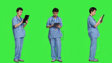 Medical assistant browsing online webpages on tablet and texting, using social media apps to chat with people. Nurse navigates internet on gadget, standing against greenscreen backdrop. Camera A.