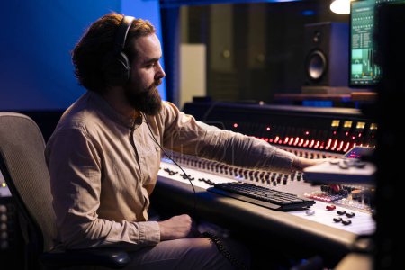 Mixer producer listening to his audio recordings and editing tracks in post production, working with technical gear and mixing console. Expert integrating amplifying sounds using motorized faders.