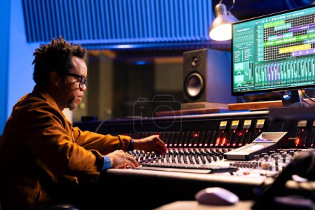 African american sound engineer works on mixing console with sliders and knobs, looking at digital software to record and process tunes. Audio technician creating new music, control room board.