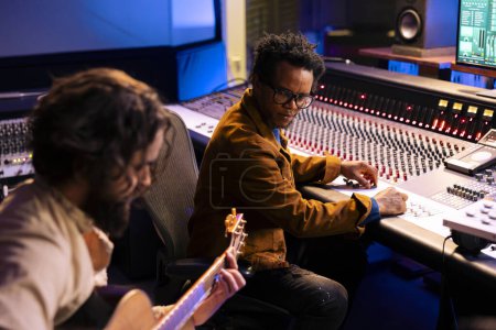 Artist playing a new record on his guitar in control room, recording and producing music with audio engineer in studio. Musician singing on electro acoustic instrument next to mixing console.