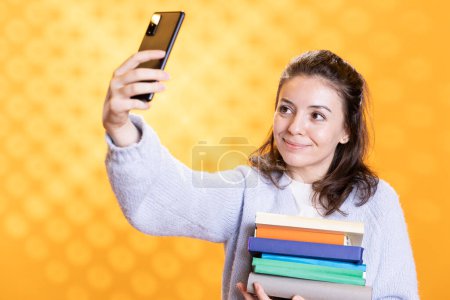 Téléchargez les photos : Femme souriante faisant selfie avec téléphone portable tenant pile de livres, isolé sur fond de studio. Étudiant heureux avec pile de manuels dans les bras prenant des photos avec téléphone portable - en image libre de droit