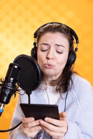 Woman frowns while doing voiceover reading of ebook on ereader to produce audiobook using dramatic acting. Narrator glowers, portraying character, recording novel using digital tablet, studio