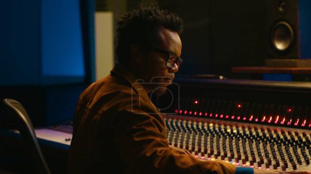 Portrait of african american audio technician uses mixing console with knobs and sliders in control room, editing recordings and adding sound effects. Skilled music producer at mixer. Camera A.