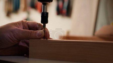 Woodworker using power drill to create holes for dowels in wooden board, close up. Carpenter sinks screws into wooden surfaces with electric tool, doing precise drilling for seamless joinery, camera A