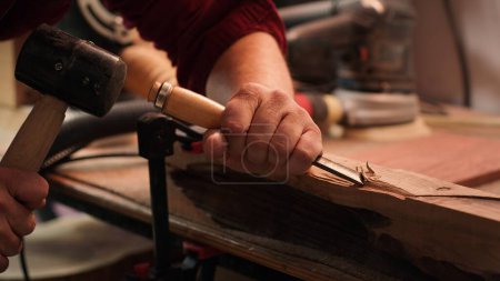 Photo for Craftsperson carving into wood using chisel and hammer in carpentry shop with precision. Manufacturer in studio shaping wooden pieces with tools, making wood art, camera A close up shot - Royalty Free Image