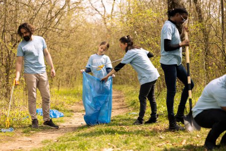 Group of diverse activists collecting trash from the forest and recycling in a garbage disposal bag, cleanup responsibility. Ecology volunteers picking up junk and plastic waste.