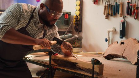 Téléchargez les photos : Sculpteur afro-américain façonnant le bois brut à l'aide d'un ciseau et d'un marteau dans une menuiserie, créant de l'art du bois, portant des lunettes de sécurité. Artiste réalisant des sculptures en bois, gravant du bois avec des outils, caméra A - en image libre de droit