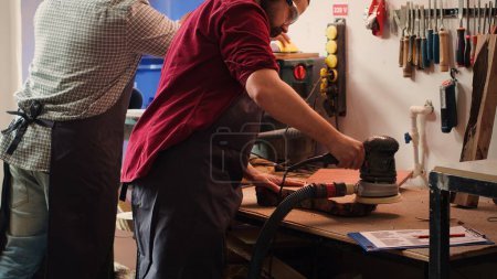 Photo for Craftsperson at work consulting blueprint and using orbital sander with sandpaper on lumber. Woodworking expert in carpentry shop uses angle grinder on wood after checking schematics, camera A - Royalty Free Image