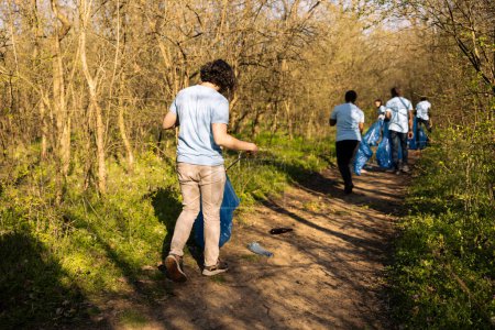 Junge Naturaktivisten sammeln mit einem langen Greifwerkzeug und Müllsack Müll ein, greifen nach Plastikmüll und Abfall, um den Wald zu säubern. Männlicher Freiwilliger leistet gemeinnützige Arbeit, globale Umweltverschmutzung.