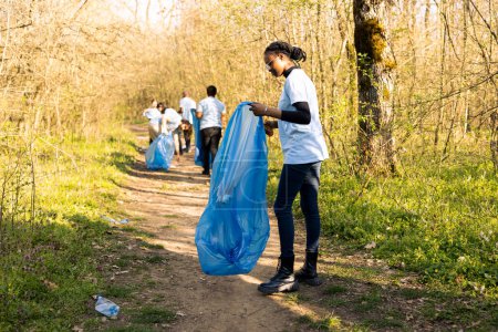 African american girl collecting garbage in a disposal blue bag, supporting nature preservation and helping protect the environment. Proud teenager volunteering to pick up trash.