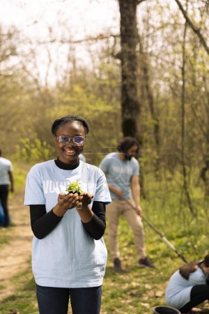 Photo for African american volunteer presents a vegetation seedling with organic soil, preserving nature and fighting pollution. Proud young girl doing voluntary work to grow trees, save the planet. - Royalty Free Image