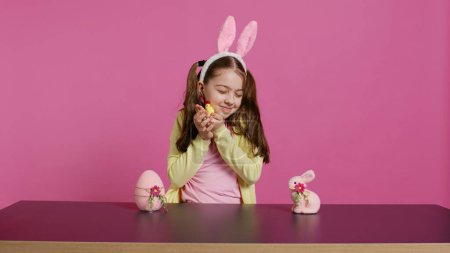 Joyful little girl playing with festive easter decorations in studio, creating arrangements with a chick, rabbit and egg. Smiling cute toddler with bunny ears showing colorful ornaments. Camera B.