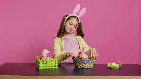 Téléchargez les photos : Jeune fille excitée arrangeant des œufs peints dans un panier pour préparer la fête de Pâques, créant des arrangements festifs. Bébé heureux ludique avec oreilles de lapin, activité créative. Caméra B. - en image libre de droit