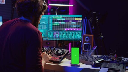 Sound engineer working on mixing and mastering techniques in his home studio, using phone with greenscreen display while he is recording audio. Artist operating soundboard and amplifier. Camera B.