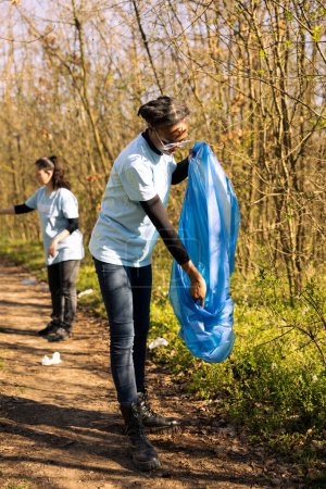 Foto de Mujer voluntaria agarrando basura y desechos plásticos con una herramienta de garra, limpiando el ecosistema natural de basura. Chica afroamericana haciendo trabajo voluntario contra el dumping ilegal. - Imagen libre de derechos