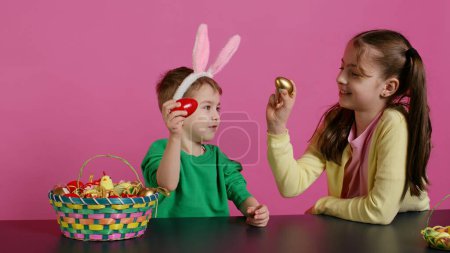 Sweet children knocking eggs together for easter tradition in studio, playing a seasonal holiday game against pink background. Lovely adorable kids having fun with festive decorations. Camera B.