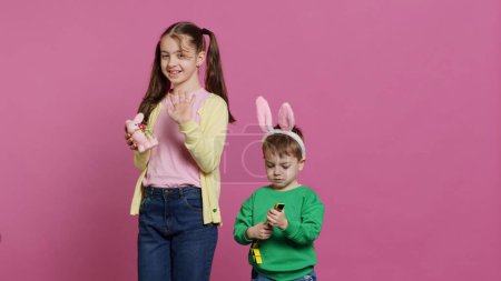 Foto de Lindo hermano y hermana posando sobre fondo rosa en el estudio, usando orejas de conejo y jugando con juguetes. Los hermanos alegres se sienten emocionados por la Pascua, las vacaciones tradicionales de primavera. Cámara A. - Imagen libre de derechos