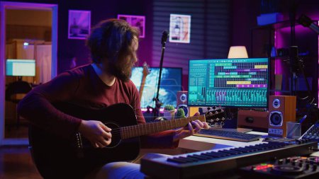 Photo for Portrait of male songwriter playing acoustic guitar and recording musical sounds to use for mix and master in home studio. Composing tunes with digital audio workstation software. Camera B. - Royalty Free Image