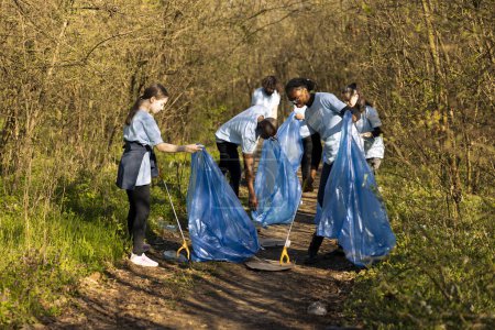 Group of diverse volunteers collecting rubbish and plastic waste, using bags to recycle and gather all junk from the forest habitat. Activists collaborating to clear the woods area from trash.