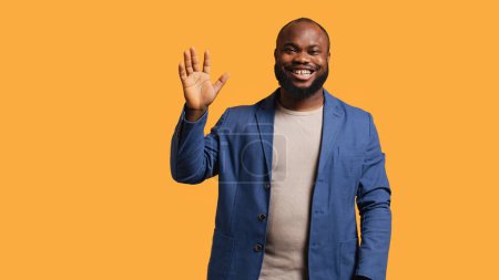Photo for African american man cheerfully doing salutation hand gesture. Portrait of happy BIPOC person raising arm to greet someone, gesturing, isolated over yellow studio background, camera A - Royalty Free Image