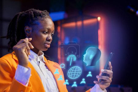 Foto de Mujer afroamericana en casa usando teléfono móvil, interactuando con asistente de inteligencia artificial usando AR. Persona que trabaja con la realidad aumentada, resolviendo tareas en Internet ayudada por la IA - Imagen libre de derechos