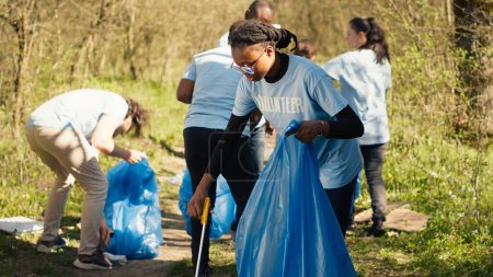 African american girl picking up trash with a long claw and garbage bags, cleaning forest habitat and fighting illegal dumping with a team of volunteers. Activist collecting rubbish. Camera A.