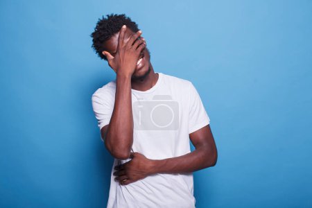Photo for Portrait of joyful young man with afro, laughing at camera while standing in studio with blue background. African american guy with his hands covering eyes and stomach is smiling and chuckling. - Royalty Free Image