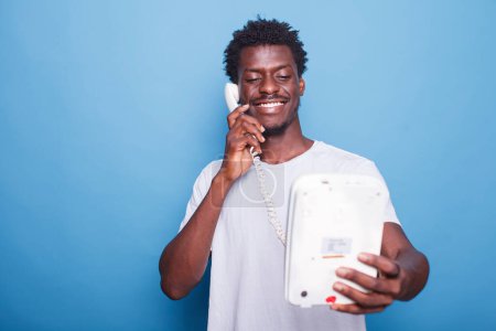 Téléchargez les photos : Un jeune homme noir debout sur un fond bleu isolé répond à un appel téléphonique. Portrait d'un homme souriant afro-américain profitant d'une conversation téléphonique fixe avec ses proches. - en image libre de droit
