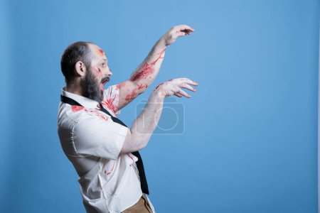 Photo for Possessed man covered in blood turned into zombie after being killed, haunting place. Wounded ghoulish cadaver limply walking towards victim, preparing for attack, studio background - Royalty Free Image