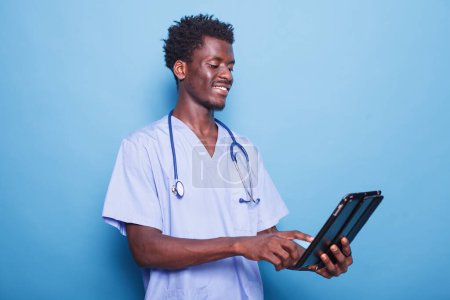 Téléchargez les photos : Smiling black doctor with stethoscope utilise une tablette pour la recherche en santé. Un professionnel de la santé afro-américain surfant sur le net sur son appareil numérique, debout sur un fond isolé. - en image libre de droit