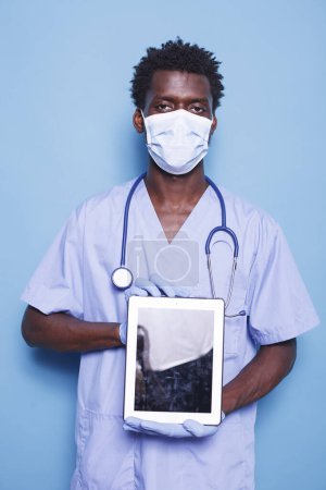 Photo for Male nurse looking at camera while holding a tablet with blank screen, against blue background. Demonstrating healthcare expertise, black man wears scrubs, face mask, gloves, and stethoscope. - Royalty Free Image