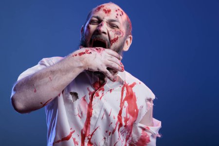 Retrato de zombi diabólico maníaco con sangre en la cara y ropa mordiéndose la mano, sintiendo hambre de carne. Loco monstruo no muerto masticando en su propio brazo, aislado sobre fondo de estudio