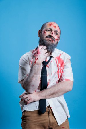 Retrato del hombre vestido de zombi ondeando por el dolor para el evento de Halloween, usando maquillaje de horror. Persona disfrazada de criatura enferma infectada cubierta de sangre y heridas, fondo del estudio