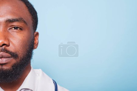 Téléchargez les photos : Gros plan d'un médecin afro-américain barbu regardant la caméra. Portrait demi-visage d'un spécialiste noir en soins de santé portant une blouse blanche sur fond bleu isolé. - en image libre de droit