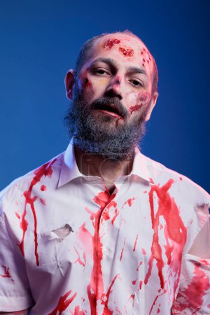 Foto de Retrato del actor interpretando a un personaje zombi en una película de terror con sangre en la cara y la ropa. Hombre vestido como criatura no muerta para la próxima película de suspenso, usando maquillaje de heridas falsas, fondo de estudio - Imagen libre de derechos