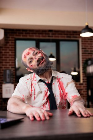 Weary man sunken on desk chair, feeling like being worked to death, making grimaces. Lifeless businessman covered in bruises and turned into zombie, concept of capitalism pressure