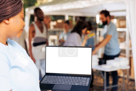 Imagen detallada que muestra a una voluntaria afroamericana llevando un portátil con una pantalla aislada de espacio de copia para personalización. Mujer negra sosteniendo minicomputador con plantilla de maqueta de pantalla blanca.