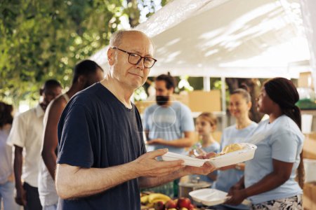 Image showcasing old caucasian homeless man holding his meal from the charitable organization. Group of volunteers at an outdoor food bank helping and feeding the hungry poor and less privileged.