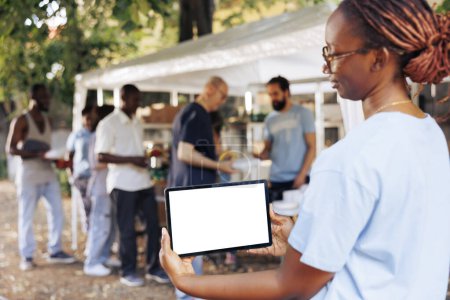 Una plantilla de maqueta aislada para mensajes filantrópicos es sostenida por una joven mujer negra sosteniendo una tableta digital. Mujer afroamericana voluntaria sosteniendo un dispositivo inteligente con una pantalla blanca en blanco.